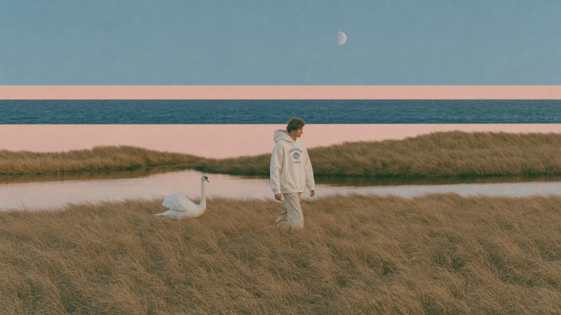 A figure in a natural Sagaponack hoodie walking through dune grass beside a swan, the ocean striped pink and navy behind